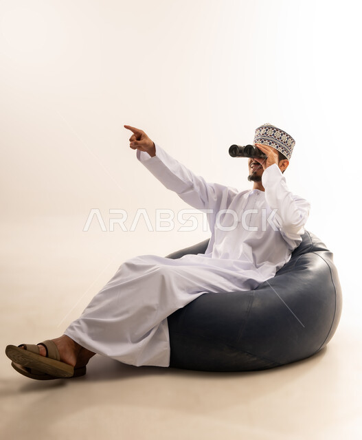 حب الاستكشاف و المغامرة, رؤية الاشياء البعيدة و تقريبها, بورتريه لشاب عربي خليجي عماني مبتسم يرتدي الدشداشة و الكمه يجلس على اريكه مريحه من الجلد الفاخر يحمل بيده منظار ثنائي العينين و ينظر الى شيء ما بإيماءات السرور والاستمتاع، خلفية بيضاء