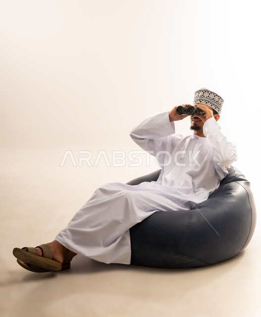 حب الاستكشاف و المغامرة, رؤية الاشياء البعيدة و تقريبها, بورتريه لشاب عربي خليجي عماني مبتسم يرتدي الدشداشة و الكمه يجلس على اريكه مريحه من الجلد الفاخر يحمل بيده منظار ثنائي العينين و ينظر الى شيء ما بإيماءات السرور والاستمتاع، خلفية بيضاء