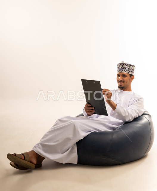 Expressions of thinking for decision making, taking notes, portrait of an Omani Arab Gulf man wearing a dishdasha and a kuma holding a notebook and pen in his hands writing information with gestures of optimism and happiness, white background