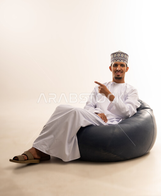 حركات يدين تدل على هذا المكان (هنا)، تحديد موقع شيء ما، بورتريه لرجل عربي خليجي عماني مبتسم يرتدي الدشداشة والكمه يجلس على أريكه مريحه من الجلد يشير بأصبع السبابه الى اتجاه ما, خلفية بيضاء