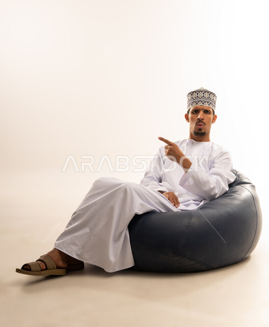 حركات يدين تدل على هذا المكان (هنا)، تحديد موقع شيء ما، بورتريه لرجل عربي خليجي عماني مبتسم يرتدي الدشداشة والكمه يجلس على أريكه مريحه من الجلد يشير بأصبع السبابه الى اتجاه ما, خلفية بيضاء