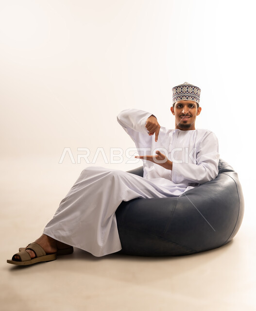 Hand gestures indicating this place (here), locating something, portrait of a smiling Omani Gulf Arab man wearing a dishdasha and a sleeve sitting on a comfortable leather sofa pointing his index finger towards his palm, white background
