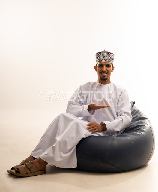 حركات يدين تدل على هذا المكان (هنا)، تحديد موقع شيء ما، بورتريه لرجل عربي خليجي عماني مبتسم يرتدي الدشداشة والكمه يجلس على أريكه مريحه من الجلد يشير بكفه الى اتجاه ما بأيماءات الترحيب والاستقبال بحفاوه, خلفية بيضاء