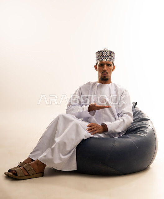 حركات يدين تدل على هذا المكان (هنا)، تحديد موقع شيء ما، بورتريه لرجل عربي خليجي عماني مبتسم يرتدي الدشداشة والكمه يجلس على أريكه مريحه من الجلد يشير بكفه الى اتجاه ما بأيماءات الترحيب والاستقبال بحفاوه, خلفية بيضاء