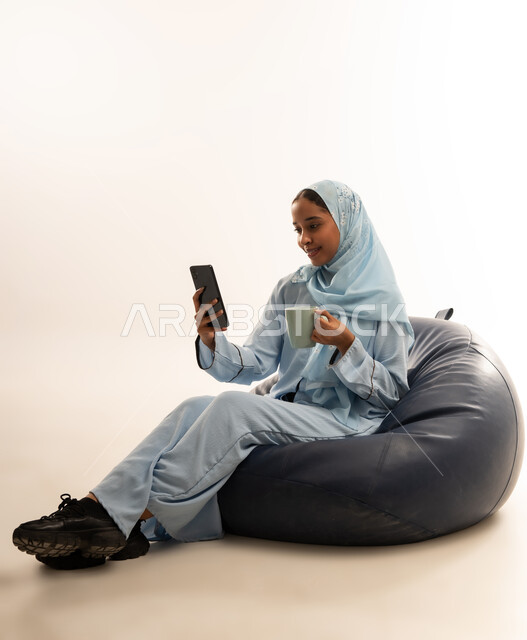 مفهوم تسهيل الاعمال والقدرة على إنجاز المهام عن بعد، استخدام التقنيات الحديثة والأجهزة المتطورة ، بورتريه لسيده عربيه خليجيه عمانيه ترتدي الحجاب والعباءه تجلس على كرسي مريح تمسك بيدها هاتفهها المحمول، خلفية بيضاء