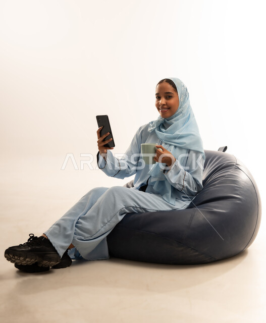 مفهوم تسهيل الاعمال والقدرة على إنجاز المهام عن بعد، استخدام التقنيات الحديثة والأجهزة المتطورة ، بورتريه لسيده عربيه خليجيه عمانيه ترتدي الحجاب والعباءه تجلس على كرسي مريح تمسك بيدها هاتفهها المحمول، خلفية بيضاء