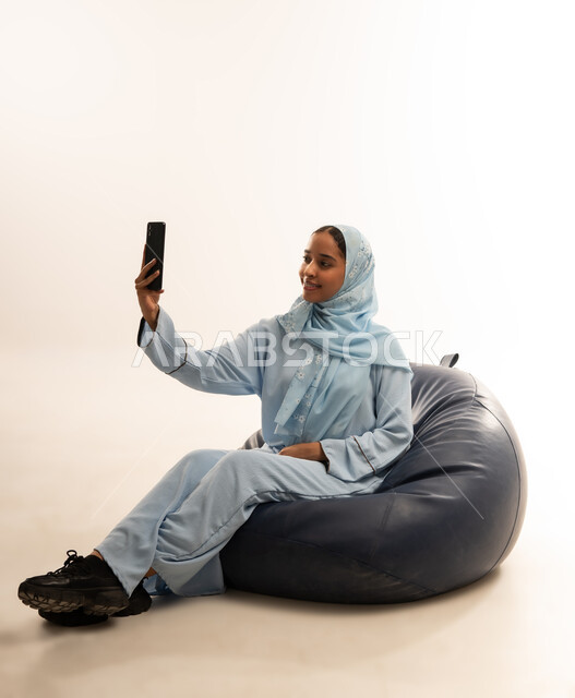 مفهوم تسهيل الاعمال والقدرة على إنجاز المهام عن بعد، استخدام التقنيات الحديثة والأجهزة المتطورة ، بورتريه لسيده عربيه خليجيه عمانيه ترتدي الحجاب والعباءه تجلس على كرسي مريح تمسك بيدها هاتفهها المحمول، خلفية بيضاء