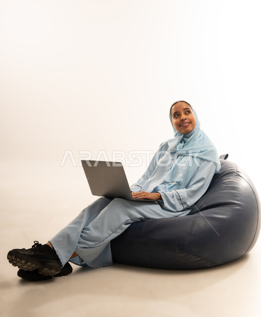 مفهوم تسهيل الاعمال والقدرة على إنجاز المهام عن بعد، استخدام التقنيات الحديثة والأجهزة المتطورة ، بورتريه لسيده عربيه خليجيه عمانيه ترتدي الحجاب والعباءه تجلس على كرسي مريح تمسك بيدها الابتوب المحمول، خلفية بيضاء