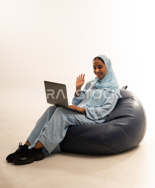 مفهوم تسهيل الاعمال والقدرة على إنجاز المهام عن بعد، استخدام التقنيات الحديثة والأجهزة المتطورة ، بورتريه لسيده عربيه خليجيه عمانيه ترتدي الحجاب والعباءه تجلس على كرسي مريح تمسك بيدها الابتوب المحمول، خلفية بيضاء