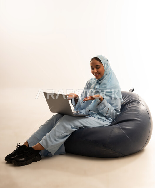 مفهوم تسهيل الاعمال والقدرة على إنجاز المهام عن بعد، استخدام التقنيات الحديثة والأجهزة المتطورة ، بورتريه لسيده عربيه خليجيه عمانيه ترتدي الحجاب والعباءه تجلس على كرسي مريح تمسك بيدها الابتوب المحمول، خلفية بيضاء