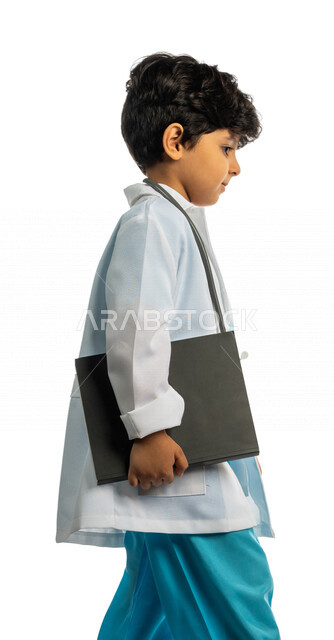 بورتريه بمنظر جانبي لفتى عربي خليجي سعودي طموح ، يرتدي زي الطبيب ، يحمل بيده كتاب وهو يمشي بإيماءات وجه ويدين مختلفة ، الطبيب الصغير ، المهنة المستقبلية ، مفهوم الدراسة وتعلم الطب ، تلميذ مجتهد ، يحلم بأن يصبح طبيب ، خلفية بيضاء