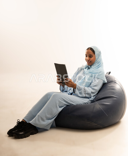 مفهوم تسهيل الاعمال والقدرة على إنجاز المهام عن بعد، استخدام التقنيات الحديثة والأجهزة المتطورة ، بورتريه لسيده عربيه خليجيه عمانيه ترتدي الحجاب والعباءه تجلس على كرسي مريح تمسك بيدها الايباد المحمول، خلفية بيضاء