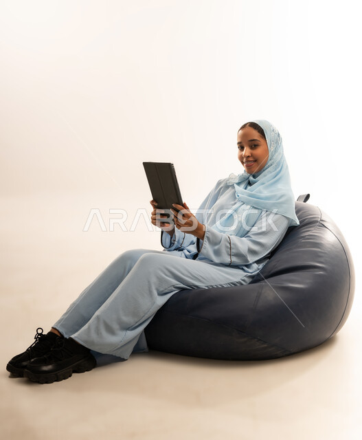 مفهوم تسهيل الاعمال والقدرة على إنجاز المهام عن بعد، استخدام التقنيات الحديثة والأجهزة المتطورة ، بورتريه لسيده عربيه خليجيه عمانيه ترتدي الحجاب والعباءه تجلس على كرسي مريح تمسك بيدها الايباد المحمول، خلفية بيضاء