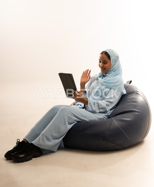 The concept of facilitating business and the ability to accomplish tasks remotely, using modern technologies and advanced devices, a portrait of an Arab Gulf Omani woman wearing a hijab and an abaya sitting on a comfortable chair holding an iPad in her hand, white background