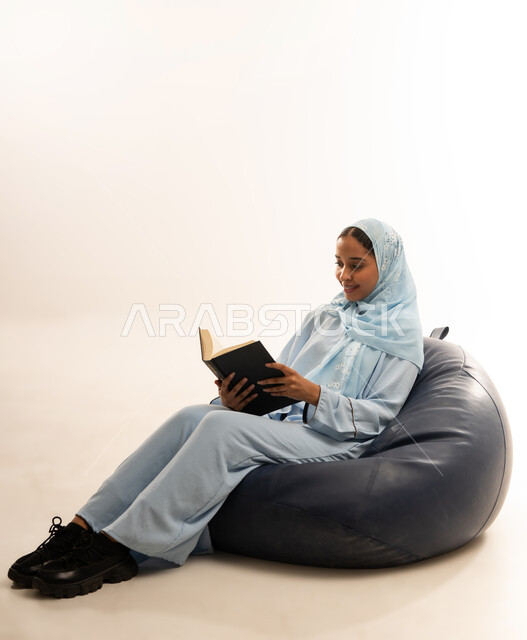 Self-learning culture, spending time reading and educating yourself, enjoying reading books, stories and novels, portrait of a veiled Omani Gulf Arab woman wearing an abaya sitting on a comfortable sofa holding a book in her hands, full body image, white background