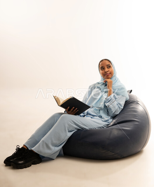 ثقافة التعلم الذاتي، قضاء الوقت بالقراءه والتثقف، الاستمتاع بالاطلاع على الكتب والقصص والروايات، بورتريه لامرأة عربية خليجية عمانية محجبة ترتدي العباءة تجلس على اريكه مريحه تحمل بيديها كتاب، صورة لكامل الجسم، خلفية بيضاء