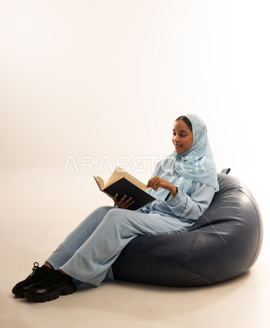ثقافة التعلم الذاتي، قضاء الوقت بالقراءه والتثقف، الاستمتاع بالاطلاع على الكتب والقصص والروايات، بورتريه لامرأة عربية خليجية عمانية محجبة ترتدي العباءة تجلس على اريكه مريحه تحمل بيديها كتاب، صورة لكامل الجسم، خلفية بيضاء