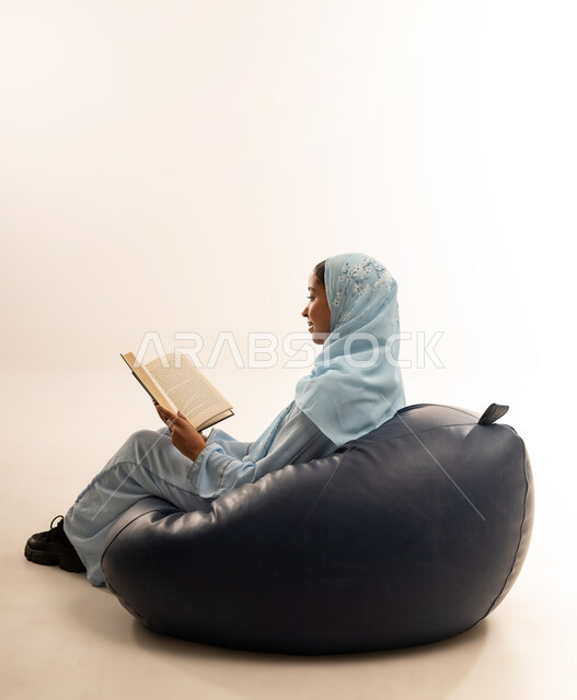 Self-learning culture, spending time reading and educating yourself, enjoying reading books, stories and novels, portrait of a veiled Omani Gulf Arab woman wearing an abaya sitting on a comfortable sofa holding a book in her hands, full body image, white background
