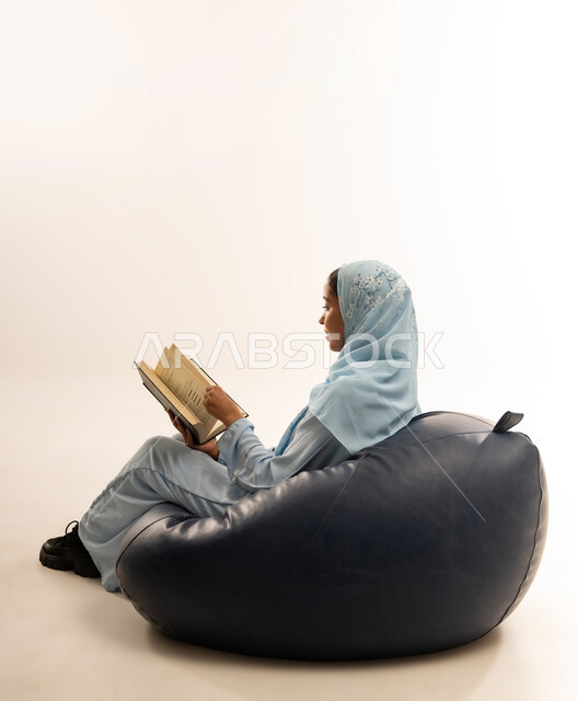 Self-learning culture, spending time reading and educating yourself, enjoying reading books, stories and novels, portrait of a veiled Omani Gulf Arab woman wearing an abaya sitting on a comfortable sofa holding a book in her hands, full body image, white background
