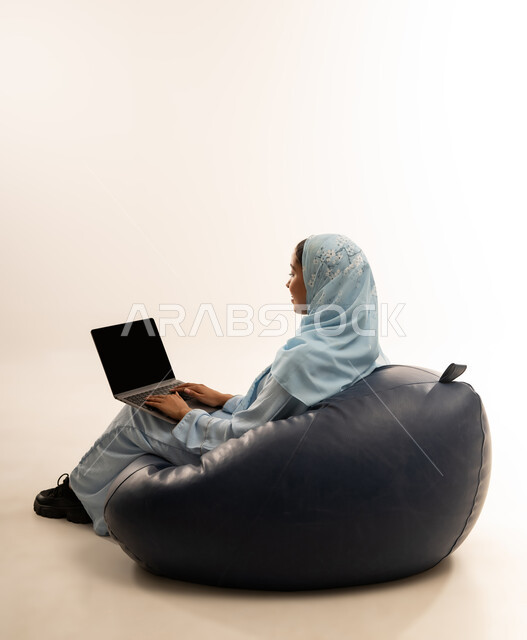 مفهوم تسهيل الاعمال والقدرة على إنجاز المهام عن بعد، استخدام التقنيات الحديثة والأجهزة المتطورة ، بورتريه لسيده عربيه خليجيه عمانيه ترتدي الحجاب والعباءه تجلس على كرسي مريح تمسك بيدها الابتوب المحمول، خلفية بيضاء