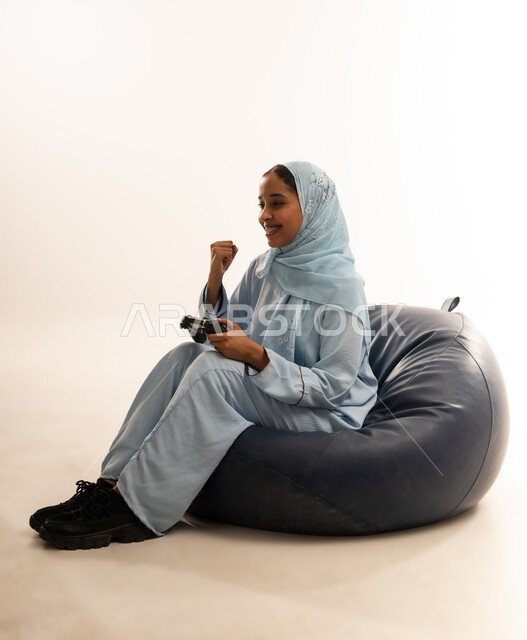 قضاء أوقات الفراغ في الفيديو غيمز، إيماءات وتعابير وجه ويدين تدل على الفوز والانتصار، بورتريه لسيده عربيه عمانيه خليجية ترتدي الحجاب والعباءة تجلس على كرسي مريح تحمل بيدها عصا التحكم وتلعب بألعاب الفيديو، خلفية بيضاء