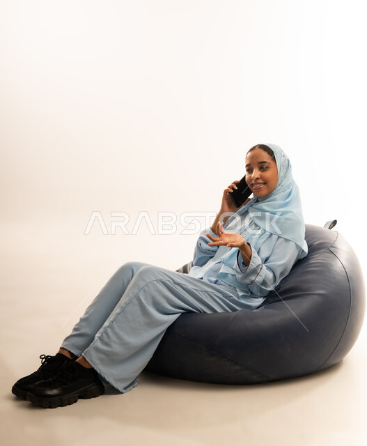الإجابة والرد على الاتصالات بإستخدام الهاتف المحمول، التواصل والاطمئنان على الأصدقاء, قضاء اوقات ممتعة بتبادل الاخبار والأحاديث، بورتريه لامرأة عربية عمانيه خليجية محجبة تجلس على أريكه مريحه من الجلد الفاخر تقوم بإجراء مكالمة صوتية عبر الجوال، خلفيه بيضاء
