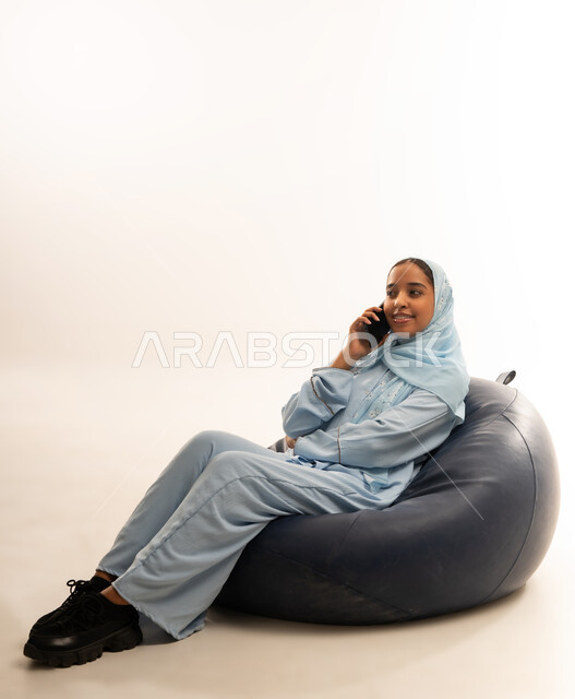 الإجابة والرد على الاتصالات بإستخدام الهاتف المحمول، التواصل والاطمئنان على الأصدقاء, قضاء اوقات ممتعة بتبادل الاخبار والأحاديث، بورتريه لامرأة عربية عمانيه خليجية محجبة تجلس على أريكه مريحه من الجلد الفاخر تقوم بإجراء مكالمة صوتية عبر الجوال، خلفيه بيضاء