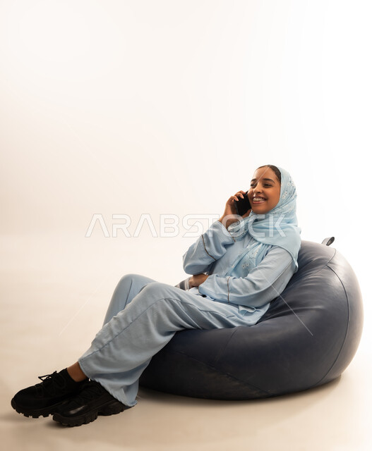 الإجابة والرد على الاتصالات بإستخدام الهاتف المحمول، التواصل والاطمئنان على الأصدقاء, قضاء اوقات ممتعة بتبادل الاخبار والأحاديث، بورتريه لامرأة عربية عمانيه خليجية محجبة تجلس على أريكه مريحه من الجلد الفاخر تقوم بإجراء مكالمة صوتية عبر الجوال، خلفيه بيضاء
