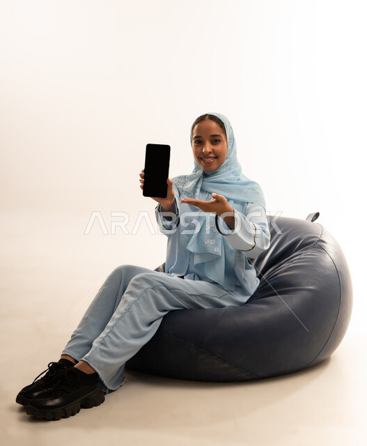 استخدام الأجهزة التقنية الحديثة للتسلية والترفيه, النظر إلى الكاميرا بإيماءات السعادة و السرور, بورتريه لشابة عربية خليجية عمانيه مبتسمة محجبة ترتدي العباءة تجلس على أريكه من الجلد تمسك بيدها الهاتف المحمول, عرض شاشة سوداء فارغة عبر الجوال, خلفية بيضاء