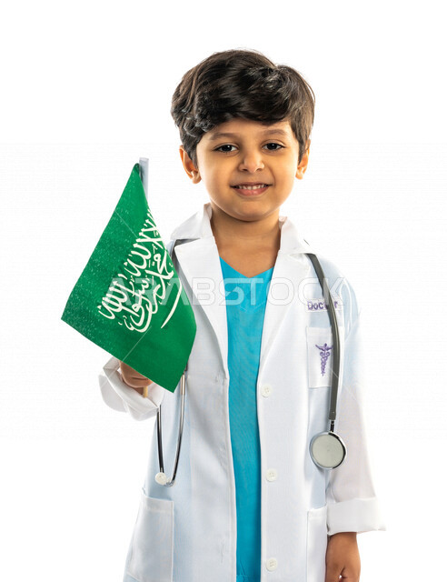 بورتريه لفتى عربي خليجي سعودي مبتسم ، يحمل بيده علم المملكة العربية السعودية ، ذكرى اليوم الوطني السعودي ، الإحتفال بالعيد الوطني السعودي ، يرتدي زي الطبيب ، الطبيب الصغير ، المهنة المستقبلية ، مفهوم الطب ، خلفية بيضاء