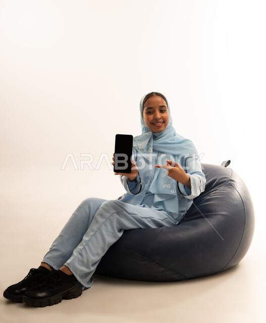Using modern technical devices for entertainment and amusement, looking at the camera with gestures of happiness and pleasure, portrait of a smiling young Arab Gulf Omani woman wearing the abaya, sitting on a leather sofa holding a mobile phone, displaying a blank black screen on the mobile, white background