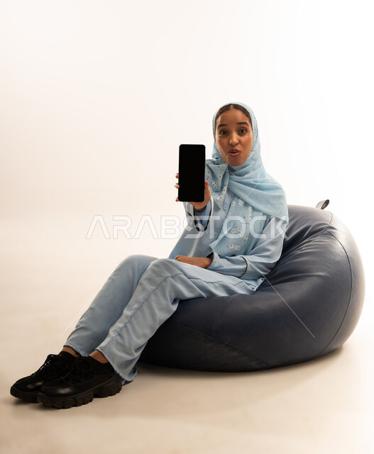 استخدام الأجهزة التقنية الحديثة للتسلية والترفيه, النظر إلى الكاميرا بإيماءات السعادة و السرور, بورتريه لشابة عربية خليجية عمانيه مبتسمة محجبة ترتدي العباءة تجلس على أريكه من الجلد تمسك بيدها الهاتف المحمول, عرض شاشة سوداء فارغة عبر الجوال, خلفية بيضاء