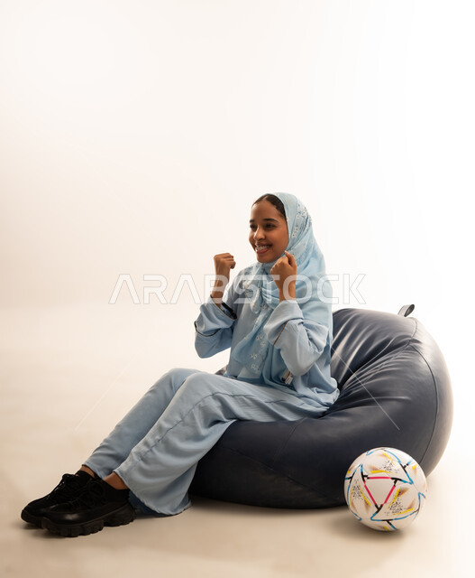 الشغف بمتابعه مباريات كرة القدم، النظر إلى الكاميرا بإيماءات التشجيع الفوز الانتصار او الخساره، بورتريه لشابة عربية عمانيه خليجية ترتدي العباءه الملونة تجلس على كرسي مريح تتابع فريقها المفضل وبجوارها كره بلاستيكيه, تشجيع الأنديه والمنتخبات، خلفية بيضاء