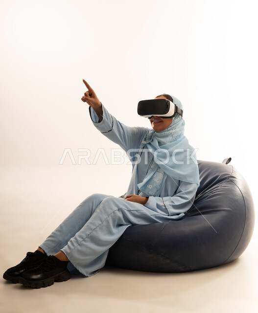 Technical and technological development, enjoying watching virtual reality movies and simulating the imaginary world, portrait of a veiled Omani Gulf Arab woman wearing a colorful abaya sitting on a comfortable chair looking through virtual reality glasses (3D VR), white background
