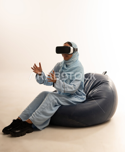 التطور التقني والتكنولوجي, الاستمتاع بمشاهدة أفلام الواقع الافتراضي ومحاكاة العالم الخيالي, بورتريه لسيده عربية خليجية عمانيه محجبة ترتدي العباءه الملونه تجلس على كرسي مريح تنظر من خلال نظارة الواقع الافتراضي (في آر ثلاثية الأبعاد), خلفية بيضاء