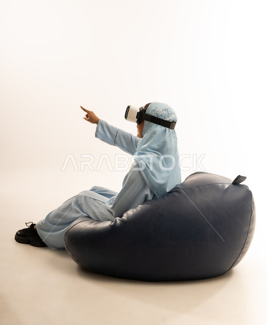 التطور التقني والتكنولوجي, الاستمتاع بمشاهدة أفلام الواقع الافتراضي ومحاكاة العالم الخيالي, بورتريه لسيده عربية خليجية عمانيه محجبة ترتدي العباءه الملونه تجلس على كرسي مريح تنظر من خلال نظارة الواقع الافتراضي (في آر ثلاثية الأبعاد), خلفية بيضاء