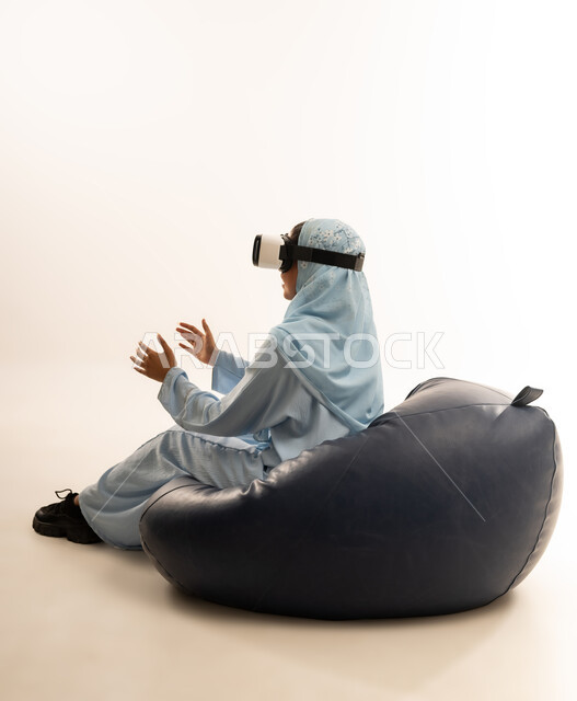 التطور التقني والتكنولوجي, الاستمتاع بمشاهدة أفلام الواقع الافتراضي ومحاكاة العالم الخيالي, بورتريه لسيده عربية خليجية عمانيه محجبة ترتدي العباءه الملونه تجلس على كرسي مريح تنظر من خلال نظارة الواقع الافتراضي (في آر ثلاثية الأبعاد), خلفية بيضاء