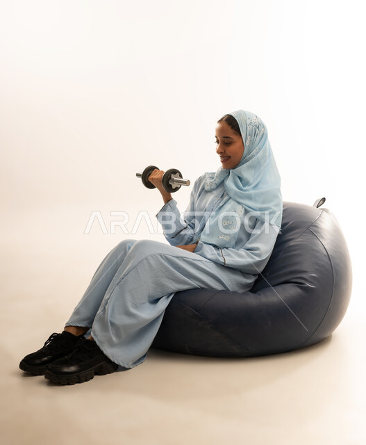 Exercising, a sporting activity to maintain a healthy body, a portrait of an Arab Gulf Omani girl wearing a colorful hijab and abaya sitting on a luxurious leather chair doing weightlifting exercises (dumbbells), strengthening and toning the arm muscles, white background
