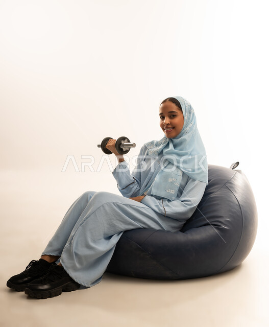 ممارسة التمارين الرياضية، نشاط رياضي للمحافظة على صحة الجسم، بورتريه  لفتاة عربية خليجية عمانيه ترتدي الحجاب والعباءه الملونه تجلس على كرسي من الجلد الفاخر تقوم بتمارين رفع الأثقال (دمبلز)، تقويه وشد عضلات الذراعيين، خلفية بيضاء