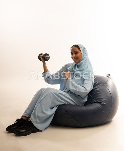 ممارسة التمارين الرياضية، نشاط رياضي للمحافظة على صحة الجسم، بورتريه  لفتاة عربية خليجية عمانيه ترتدي الحجاب والعباءه الملونه تجلس على كرسي من الجلد الفاخر تقوم بتمارين رفع الأثقال (دمبلز)، تقويه وشد عضلات الذراعيين، خلفية بيضاء