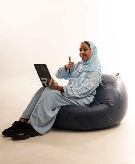 التسلية والترفيه وقضاء أوقات ممتعه, استخدام التكنولوجيا و الأجهزة التقنية الحديثة المتطورة, بورتريه لفتاة عربية خليجية عمانيه محجبة ترتدي العباءة تجلس على كرسي مريح تحمل بيدها الايباد تشير اليه بايماءات السعادة والسرور, خلفية بيضاء