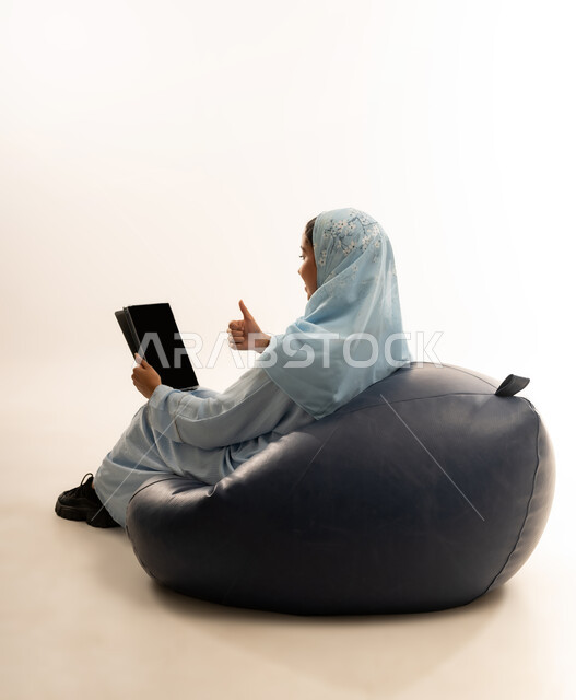 Entertainment and fun, using modern technology and advanced technical devices, portrait of a veiled Omani Gulf Arab girl wearing an abaya sitting on a comfortable chair holding an iPad in her hand, pointing at it with gestures of happiness and pleasure, white background