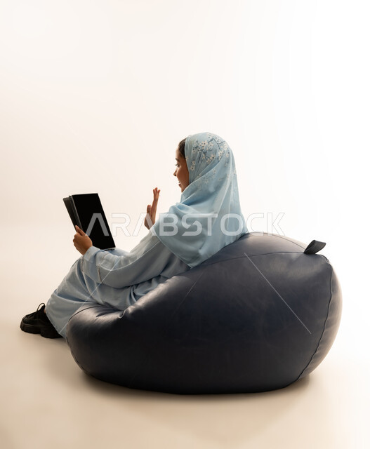 Entertainment and fun, using modern technology and advanced technical devices, portrait of a veiled Omani Gulf Arab girl wearing an abaya sitting on a comfortable chair holding an iPad in her hand, pointing at it with gestures of happiness and pleasure, white background