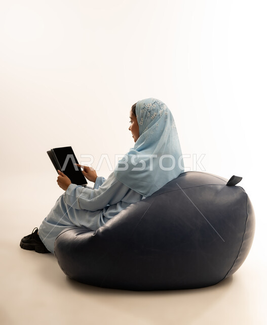 التسلية والترفيه وقضاء أوقات ممتعه, استخدام التكنولوجيا و الأجهزة التقنية الحديثة المتطورة, بورتريه لفتاة عربية خليجية عمانيه محجبة ترتدي العباءة تجلس على كرسي مريح تحمل بيدها الايباد تشير اليه بايماءات السعادة والسرور, خلفية بيضاء