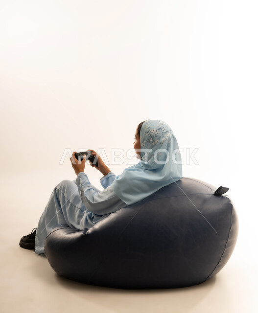 Entertaining modern technologies, portrait from the back of an Omani Gulf Arab woman sitting on a comfortable chair holding a joystick in her hand and playing video games, spending free time playing PlayStation entertainment games, white background