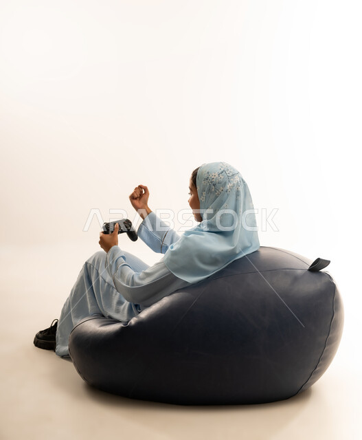 تقنيات حديثة مسلية, بورتريه من الخلف لإمرأة عربية عمانيه خليجية تجلس على كرسي مريح تحمل بيدها عصا التحكم و تلعب بألعاب الفيديو ، قضاء أوقات الفراغ باللعب بألعاب البلايستيشن الترفيهية ، خلفية بيضاء