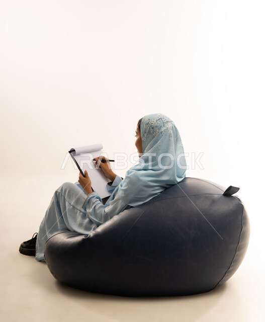 Writing and taking notes, contemplation and concentration, gestures indicating thinking, portrait of an Arab Gulf Omani woman wearing a colorful abaya sitting on a comfortable chair holding a tablet and pen, white background