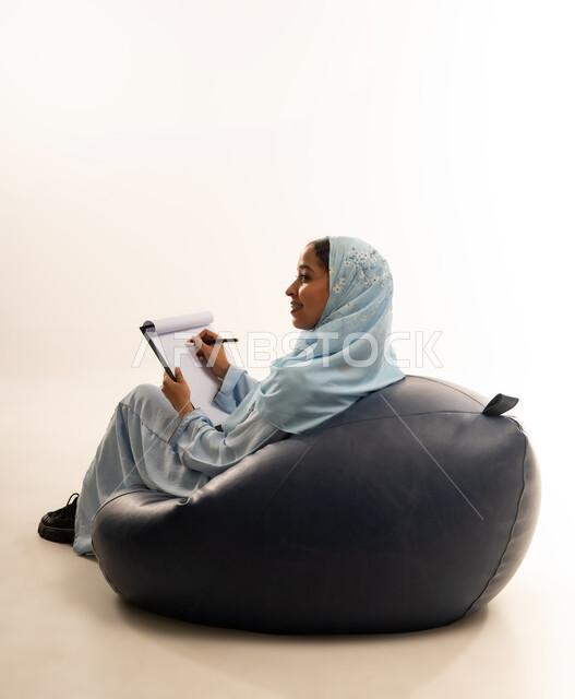 Writing and taking notes, contemplation and concentration, gestures indicating thinking, portrait of an Arab Gulf Omani woman wearing a colorful abaya sitting on a comfortable chair holding a tablet and pen, white background