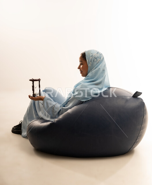 الجلوس بتعابير الملل والانتظار والسؤال, إدارة الأعمال وتنظيم الجدول الزمني, القيام بمراقبة توقيت الرمل, إدراك قيمة اللحظات الثمينة, بورتريه لشابة عربية خليجية عمانيه ترتدي العباءة الملونة والحجاب تمسك بيدها الساعة الرملية الزجاجية (البنكام), خلفية بيضاء