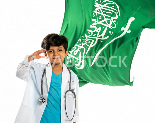 بورتريه لفتى عربي خليجي سعودي مبتسم ، يلقي التحية لوطنه الحبيب المملكة العربية السعودية ، خلفه علم المملكة ، ذكرى اليوم الوطني السعودي ، الإحتفال بالعيد الوطني السعودي ، يرتدي زي الطبيب ، الطبيب الصغير ، المهنة المستقبلية ، مفهوم الطب ، خلفية بيضاء
